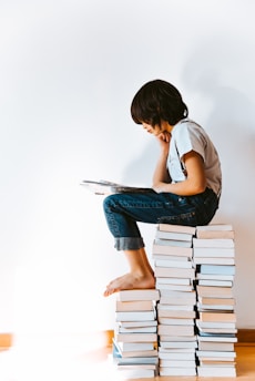 A young boy sits atop two large stacks of books, deeply engrossed in reading. He has a thoughtful expression, resting his chin on one hand, while his bare feet dangle. The background is a plain, light-colored wall that enhances the focus on him and the books.