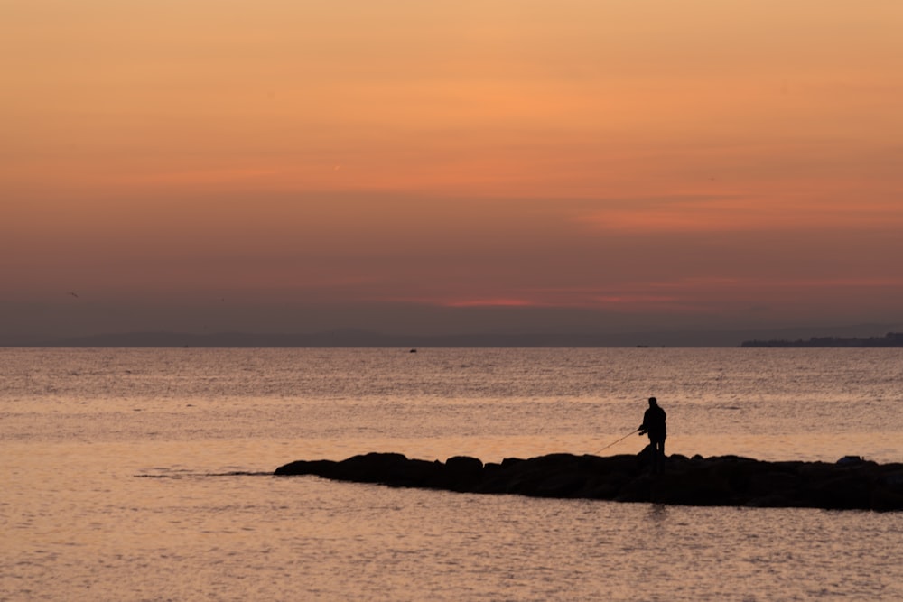 silhouette of person sitting on rock near body of water during sunset