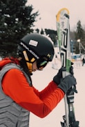 A person dressed in winter sports gear is preparing for skiing. They are wearing a black helmet with stickers, goggles, a red jacket, and gray gloves. The individual is holding a pair of skis upright while standing on a snowy landscape. In the background, there are trees and ski equipment racks visible.