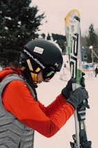 A person dressed in winter sports gear is preparing for skiing. They are wearing a black helmet with stickers, goggles, a red jacket, and gray gloves. The individual is holding a pair of skis upright while standing on a snowy landscape. In the background, there are trees and ski equipment racks visible.