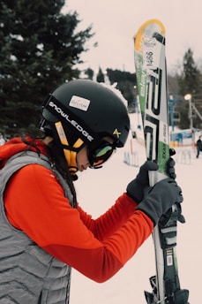 A person dressed in winter sports gear is preparing for skiing. They are wearing a black helmet with stickers, goggles, a red jacket, and gray gloves. The individual is holding a pair of skis upright while standing on a snowy landscape. In the background, there are trees and ski equipment racks visible.