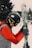 A person dressed in winter sports gear is preparing for skiing. They are wearing a black helmet with stickers, goggles, a red jacket, and gray gloves. The individual is holding a pair of skis upright while standing on a snowy landscape. In the background, there are trees and ski equipment racks visible.