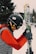 A person dressed in winter sports gear is preparing for skiing. They are wearing a black helmet with stickers, goggles, a red jacket, and gray gloves. The individual is holding a pair of skis upright while standing on a snowy landscape. In the background, there are trees and ski equipment racks visible.