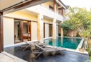 A modern outdoor patio area featuring a private swimming pool with clear blue water. Wooden deck chairs with towels are placed near the pool, offering a relaxing atmosphere. The area is surrounded by lush greenery and tropical plants, adding a touch of nature. Large glass doors behind the pool open into a well-appointed room.