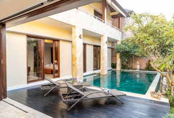 A modern outdoor patio area featuring a private swimming pool with clear blue water. Wooden deck chairs with towels are placed near the pool, offering a relaxing atmosphere. The area is surrounded by lush greenery and tropical plants, adding a touch of nature. Large glass doors behind the pool open into a well-appointed room.