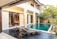 A modern outdoor patio area featuring a private swimming pool with clear blue water. Wooden deck chairs with towels are placed near the pool, offering a relaxing atmosphere. The area is surrounded by lush greenery and tropical plants, adding a touch of nature. Large glass doors behind the pool open into a well-appointed room.