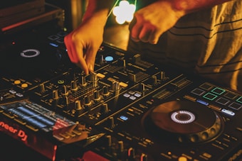 A person manipulating a DJ controller with various faders, knobs, and buttons in a dimly lit setting, creating an atmosphere of a lively music event.