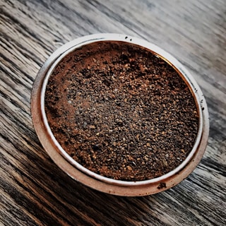 A metallic portafilter filled with finely ground coffee sits on a textured wooden surface. The ground coffee is dark brown and evenly distributed across the surface of the portafilter.