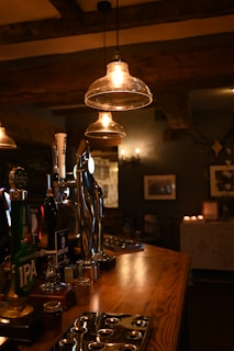 Warm hanging lights illuminate a rustic bar area with several beer taps, including brands such as IPA. The wooden bar counter adds a cozy feel, complemented by dim lighting and a few small candles in the background. Framed pictures hang on the dark walls, enhancing the vintage ambiance.