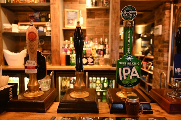 A row of traditional beer taps is lined up on a wooden bar inside a cozy pub setting, with various types of beer and ale bottles displayed on shelves in the background. Warm lighting creates a rustic and inviting atmosphere.