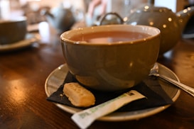 A large cup filled with tea rests on a saucer placed on a wooden table. The saucer holds a small biscuit and a wrapped sugar packet alongside the cup. In the background, there are blurred images of more cups and a teapot, suggesting a cozy setting.