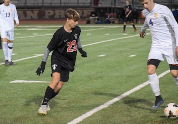 Two soccer players are captured in mid-action during a game on a grass field. One player is wearing a black jersey and shorts, marked with the number 23, and is positioned defensively. Another player in a white jersey, numbered 4, is in the process of controlling the ball with their foot. The background shows more players and part of the stands, suggesting an active and competitive environment.
