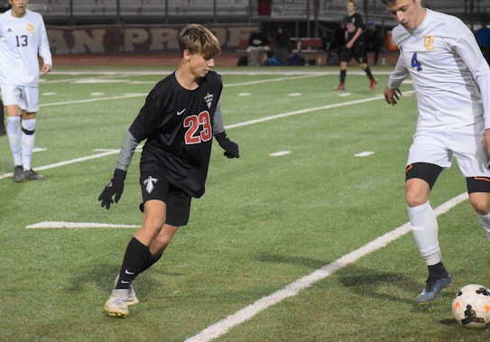 Two soccer players are captured in mid-action during a game on a grass field. One player is wearing a black jersey and shorts, marked with the number 23, and is positioned defensively. Another player in a white jersey, numbered 4, is in the process of controlling the ball with their foot. The background shows more players and part of the stands, suggesting an active and competitive environment.