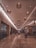 An indoor shopping mall corridor with warm lighting and hanging basket-style lamps along the ceiling. The floor is made of polished stone tiles, creating a reflective surface. Shoppers are seen walking and pushing shopping carts. Storefronts with varied signage line both sides of the passageway.
