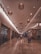 An indoor shopping mall corridor with warm lighting and hanging basket-style lamps along the ceiling. The floor is made of polished stone tiles, creating a reflective surface. Shoppers are seen walking and pushing shopping carts. Storefronts with varied signage line both sides of the passageway.