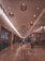 An indoor shopping mall corridor with warm lighting and hanging basket-style lamps along the ceiling. The floor is made of polished stone tiles, creating a reflective surface. Shoppers are seen walking and pushing shopping carts. Storefronts with varied signage line both sides of the passageway.