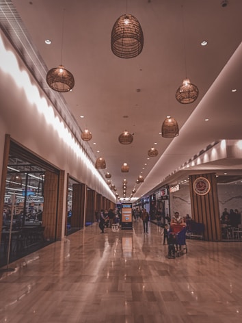 An indoor shopping mall corridor with warm lighting and hanging basket-style lamps along the ceiling. The floor is made of polished stone tiles, creating a reflective surface. Shoppers are seen walking and pushing shopping carts. Storefronts with varied signage line both sides of the passageway.
