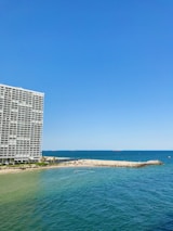 A large oceanfront building with balconies stands prominently on the left side. To the right, a sandy beach stretches out, meeting the vibrant blue of the sea. Small waves ripple across the water, and several small boats or watercraft can be seen near the shoreline. The sky is clear, showcasing a bright blue hue.