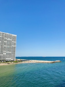 A large oceanfront building with balconies stands prominently on the left side. To the right, a sandy beach stretches out, meeting the vibrant blue of the sea. Small waves ripple across the water, and several small boats or watercraft can be seen near the shoreline. The sky is clear, showcasing a bright blue hue.