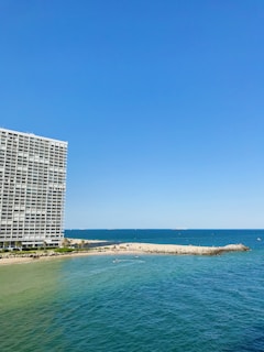 A large oceanfront building with balconies stands prominently on the left side. To the right, a sandy beach stretches out, meeting the vibrant blue of the sea. Small waves ripple across the water, and several small boats or watercraft can be seen near the shoreline. The sky is clear, showcasing a bright blue hue.