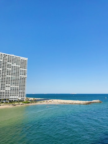 A large oceanfront building with balconies stands prominently on the left side. To the right, a sandy beach stretches out, meeting the vibrant blue of the sea. Small waves ripple across the water, and several small boats or watercraft can be seen near the shoreline. The sky is clear, showcasing a bright blue hue.