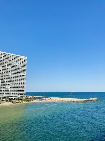 A large oceanfront building with balconies stands prominently on the left side. To the right, a sandy beach stretches out, meeting the vibrant blue of the sea. Small waves ripple across the water, and several small boats or watercraft can be seen near the shoreline. The sky is clear, showcasing a bright blue hue.
