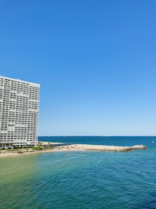 A large oceanfront building with balconies stands prominently on the left side. To the right, a sandy beach stretches out, meeting the vibrant blue of the sea. Small waves ripple across the water, and several small boats or watercraft can be seen near the shoreline. The sky is clear, showcasing a bright blue hue.