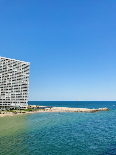 A large oceanfront building with balconies stands prominently on the left side. To the right, a sandy beach stretches out, meeting the vibrant blue of the sea. Small waves ripple across the water, and several small boats or watercraft can be seen near the shoreline. The sky is clear, showcasing a bright blue hue.