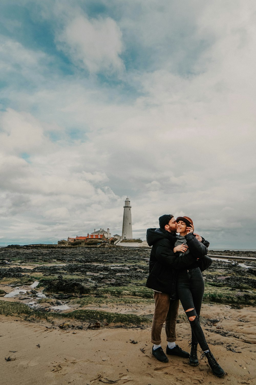 a man and woman hugging on the beach with a lighthouse in the background