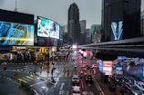 A bustling urban intersection with tall buildings and large digital billboards displaying advertisements. The streets are wet from rain, and there are several vehicles and pedestrians crossing the road. Bright lights from the billboards and streetlights illuminate the scene, creating reflections on the wet surface. The surrounding architecture includes modern skyscrapers and a prominent skybridge.