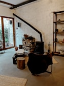 A cozy interior featuring a rustic wood-burning stove with a long black chimney pipe against a textured white wall. A stack of chopped firewood is neatly arranged next to it, with baskets and tools for tending the fire. A chair draped with a dark fur throw sits nearby, along with a small wooden stool and a low table. The room has large windows on one side, allowing natural light to illuminate the space. The decor includes wooden shelves with various items, and a rug on the floor adds warmth to the atmosphere.