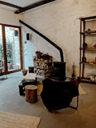 A cozy interior featuring a rustic wood-burning stove with a long black chimney pipe against a textured white wall. A stack of chopped firewood is neatly arranged next to it, with baskets and tools for tending the fire. A chair draped with a dark fur throw sits nearby, along with a small wooden stool and a low table. The room has large windows on one side, allowing natural light to illuminate the space. The decor includes wooden shelves with various items, and a rug on the floor adds warmth to the atmosphere.
