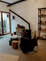 A cozy interior featuring a rustic wood-burning stove with a long black chimney pipe against a textured white wall. A stack of chopped firewood is neatly arranged next to it, with baskets and tools for tending the fire. A chair draped with a dark fur throw sits nearby, along with a small wooden stool and a low table. The room has large windows on one side, allowing natural light to illuminate the space. The decor includes wooden shelves with various items, and a rug on the floor adds warmth to the atmosphere.