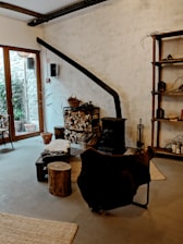 A cozy interior featuring a rustic wood-burning stove with a long black chimney pipe against a textured white wall. A stack of chopped firewood is neatly arranged next to it, with baskets and tools for tending the fire. A chair draped with a dark fur throw sits nearby, along with a small wooden stool and a low table. The room has large windows on one side, allowing natural light to illuminate the space. The decor includes wooden shelves with various items, and a rug on the floor adds warmth to the atmosphere.