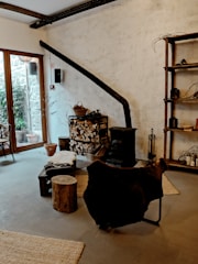 A cozy interior featuring a rustic wood-burning stove with a long black chimney pipe against a textured white wall. A stack of chopped firewood is neatly arranged next to it, with baskets and tools for tending the fire. A chair draped with a dark fur throw sits nearby, along with a small wooden stool and a low table. The room has large windows on one side, allowing natural light to illuminate the space. The decor includes wooden shelves with various items, and a rug on the floor adds warmth to the atmosphere.