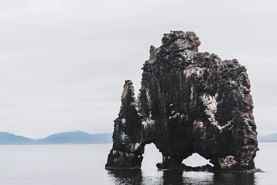 The image depicts a large, dark gray rock formation with multiple layers and crevices, situated on the shoreline of a body of water. The rock formation is surrounded by calm waters that extend into the distance, creating a serene atmosphere. In the background, there are mountains visible in the distance, adding depth to the scene.