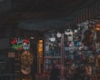 A dimly lit market stall filled with colorful and various merchandise, including boxes of different products, hanging decorations, and beauty items. There are people visible near the stall, suggesting a busy market environment.
