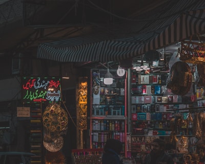 A dimly lit market stall filled with colorful and various merchandise, including boxes of different products, hanging decorations, and beauty items. There are people visible near the stall, suggesting a busy market environment.