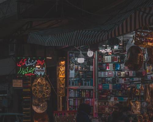 A dimly lit market stall filled with colorful and various merchandise, including boxes of different products, hanging decorations, and beauty items. There are people visible near the stall, suggesting a busy market environment.