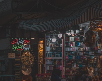 A dimly lit market stall filled with colorful and various merchandise, including boxes of different products, hanging decorations, and beauty items. There are people visible near the stall, suggesting a busy market environment.