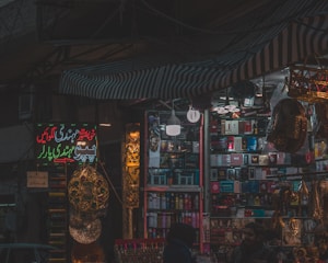 A dimly lit market stall filled with colorful and various merchandise, including boxes of different products, hanging decorations, and beauty items. There are people visible near the stall, suggesting a busy market environment.