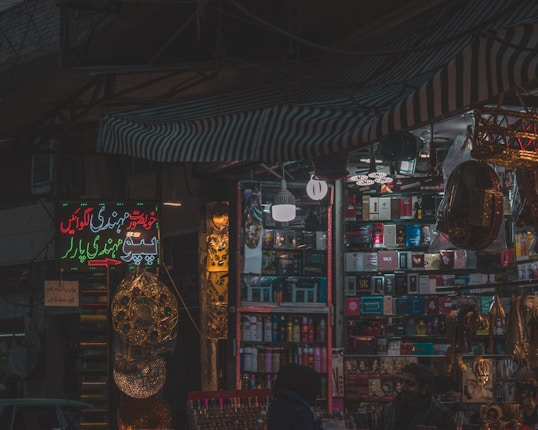 A dimly lit market stall filled with colorful and various merchandise, including boxes of different products, hanging decorations, and beauty items. There are people visible near the stall, suggesting a busy market environment.