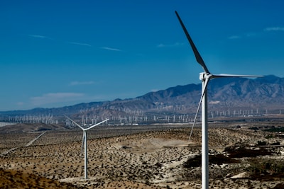 A landscape featuring a desert terrain with multiple wind turbines scattered across the valley. The scene includes a few large wind turbines in the foreground and a vast array of smaller ones stretching towards the distant mountains under a clear blue sky.