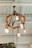 A rustic chandelier made of thick rope and Edison bulbs hangs from a ceiling with exposed beams. The design gives a warm and vintage feel, and a small amount of greenery is visible below, suggesting a cozy, natural setting.