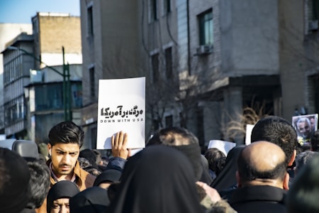 A group of people gathered in a street, holding signs including one that reads 'Down with USA'. The crowd is dressed mostly in dark colors and the background consists of buildings. A man with dark hair is prominently visible at the front of the crowd.