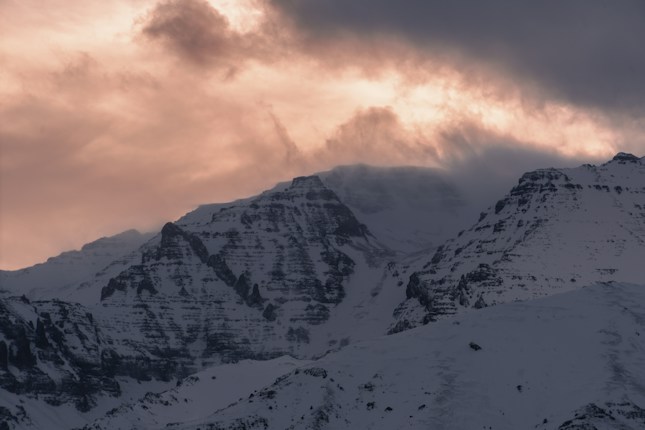 A dramatic mountain landscape at dusk with snow-covered peaks partially shrouded by dark, swirling clouds. The sky is illuminated with a warm glow, creating a striking contrast against the cold, rugged terrain.