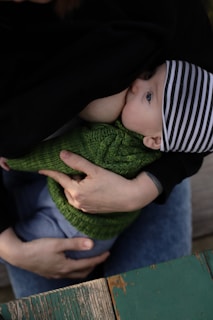 A baby wearing a striped hat and green sweater is being breastfed. The scene is intimate and tender, with the baby's eyes open while nursing. The setting includes a wooden surface and hands gently holding the infant.