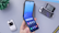 A hand is holding a modern foldable smartphone displaying a home screen with app icons and a weather widget. The phone is partially folded, and the angle reveals its flexible screen feature. In the background, two older flip and slide phones are placed on a light grey surface along with a green plant.