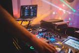 A DJ setup with a mixing console and a laptop displaying music software. Colorful lights create a vibrant atmosphere, and a DJ's hand is adjusting the controls.