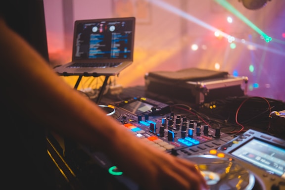 A DJ setup with a mixing console and a laptop displaying music software. Colorful lights create a vibrant atmosphere, and a DJ's hand is adjusting the controls.